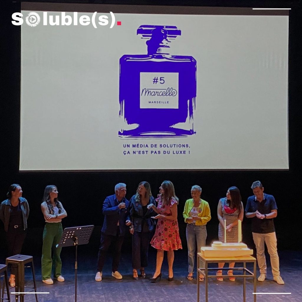 L'équipe de Marcelle en présentation sur une scène de théâtre. Sur un écran géant une publicité inspirée du flacon de parfum Chanel n°5 les surplombe. On peu lire en lettre bleu "Marcelle #5 Un média de solutions ce n'est pas du luxe !"